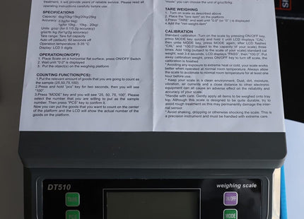 Γκρι ζυγαριά DT510 με το εγχειρίδιο χρήσης επάνω στην πλατφόρμα και ορατά τα πλήκτρα TARE, PCS, ON/OFF και MODE.